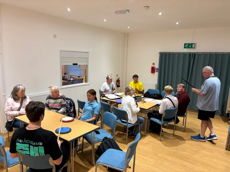 Band members of all ages at tables during a rehearsal evening, an instructor standing at the front