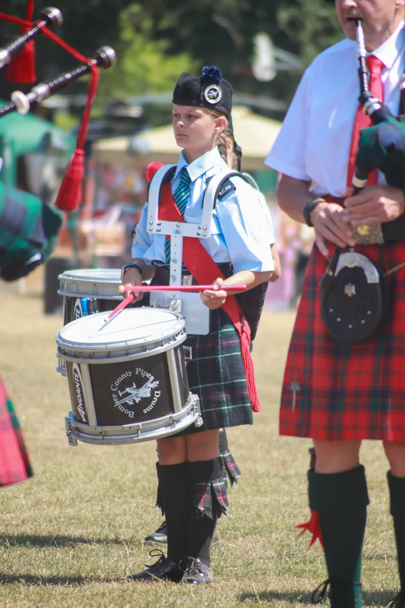 A young drummer in band uniform playing a snare drum beside a piper on a showground